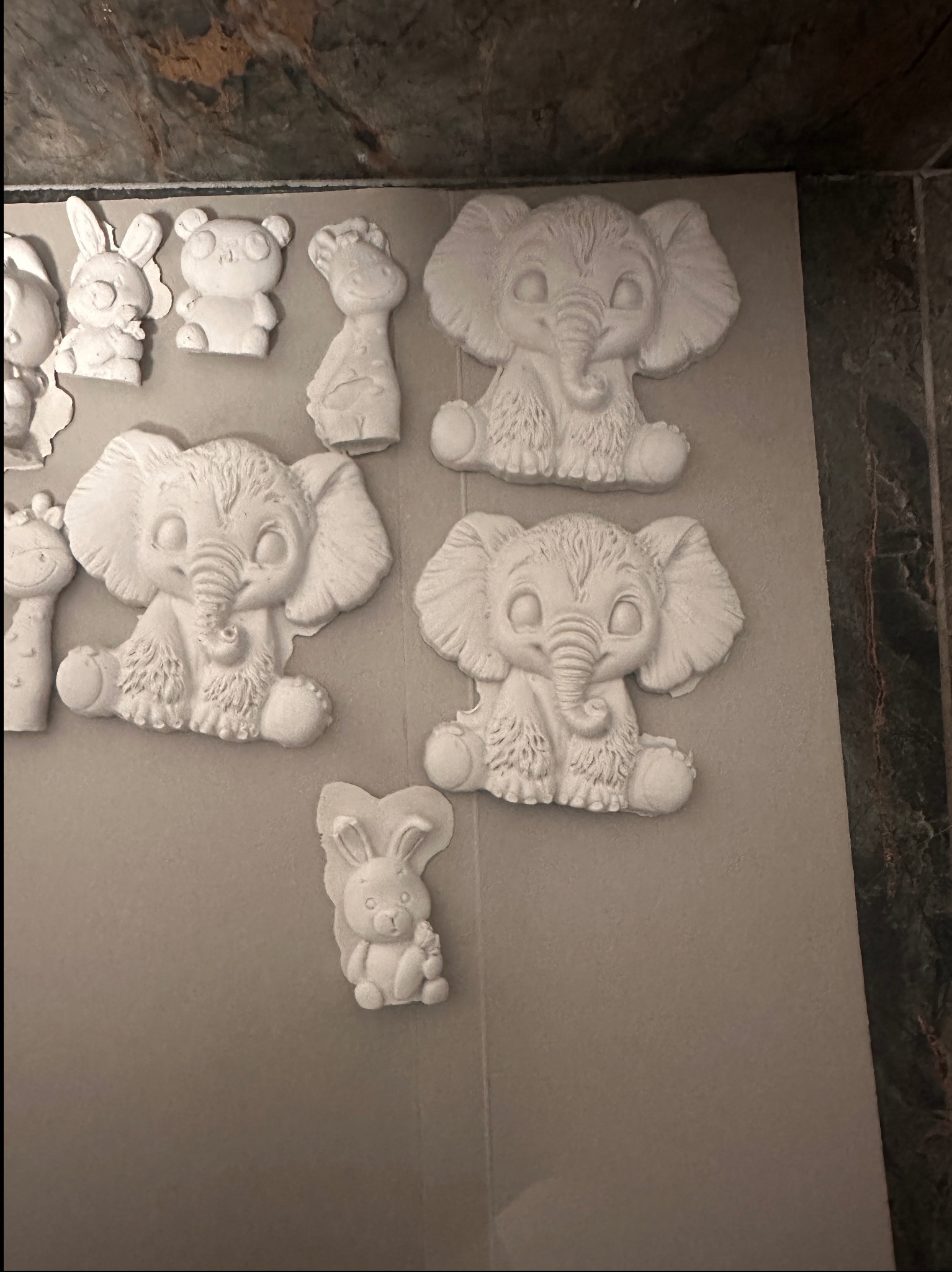 Gipsowe figurki zwierząt (słoń, królik, żyrafa, miś) na szarym tle. Ręcznie robione ozdoby dla dzieci, gotowe do malowania. Kreatywne warsztaty plastyczne.