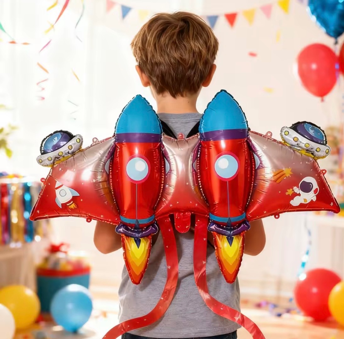 Chłopiec z tyłu, ubrany w kosmiczne skrzydła z balonów w kształcie rakiet i UFO, na tle dekoracji imprezowych i kolorowych balonów.