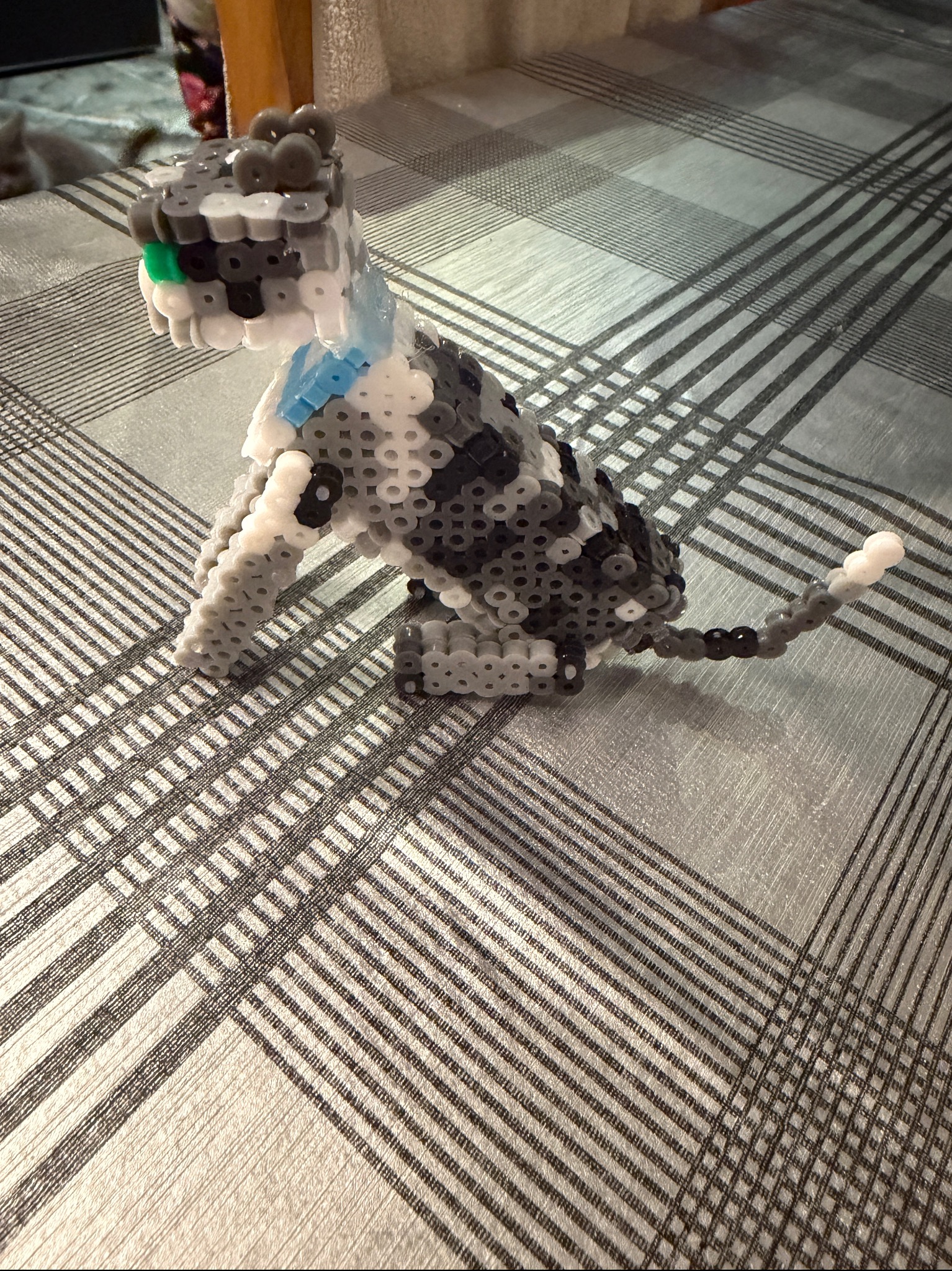 Figurka psa z koralików Hama na wzorzystym tle. Szaro-biała kolorystyka, widoczny detal zielonego koralika w oku. Rękodzieło, unikalna ozdoba.