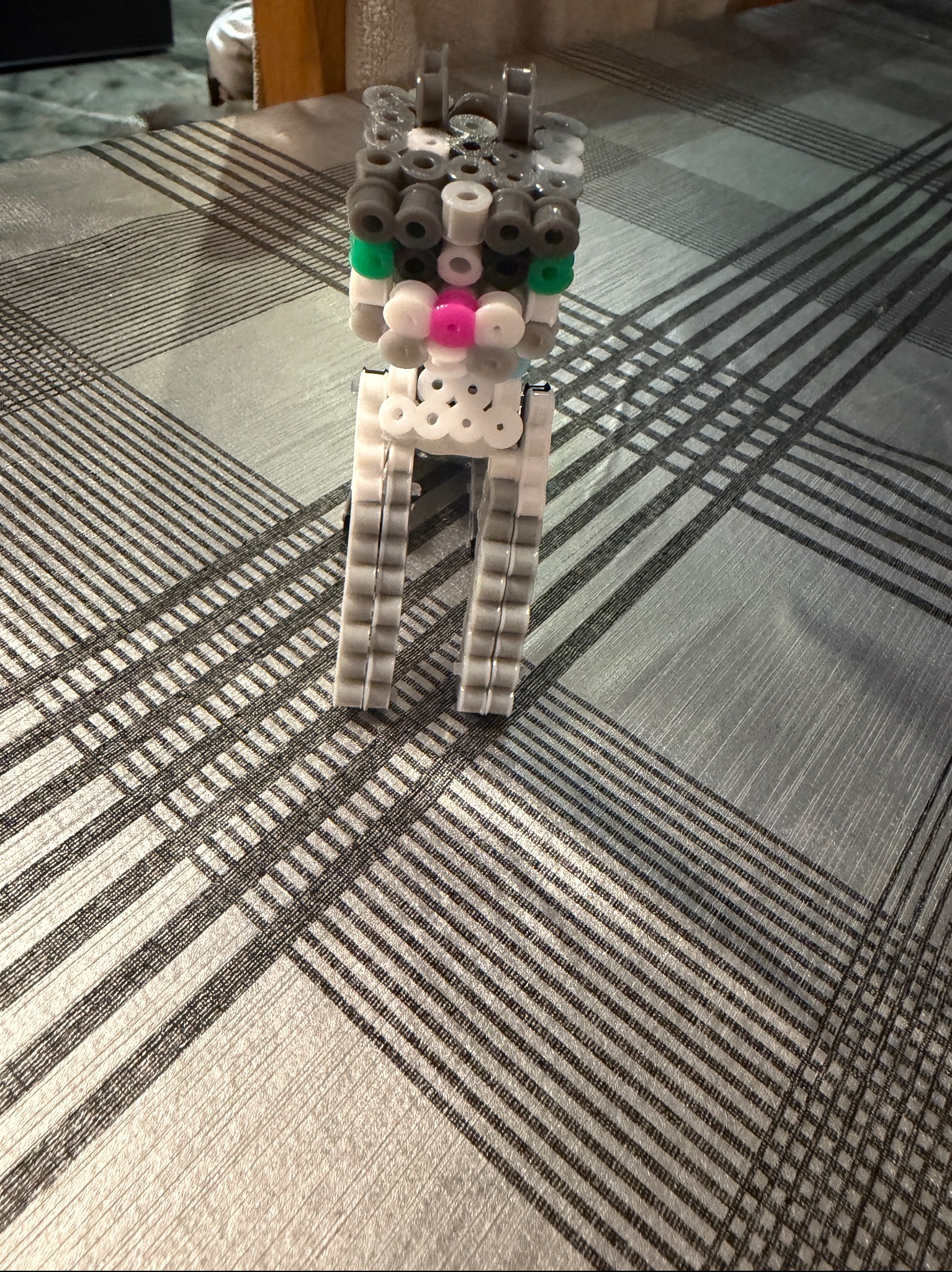 Figurka zwierzęcia, prawdopodobnie kota, wykonana z kolorowych, plastikowych koralików na tle szarej, wzorzystej powierzchni. Widoczne detale konstrukcji i użyte kolory.