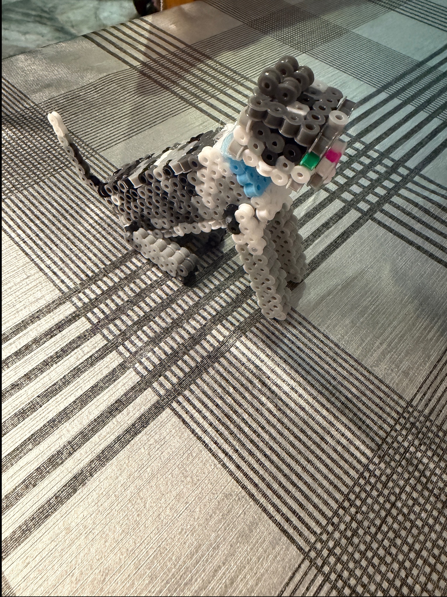 Figurka kota wykonana z kolorowych koralików Hama, stojąca na wzorzystej tkaninie. Kot ma szaro-białe umaszczenie i niebieską kokardę. Widoczne detale wykonania.