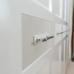 Biała szafa wnękowa z frezowanymi frontami i chromowanymi uchwytami, widok z boku, z zasłonami w tle. Detal wykończenia i elegancja w projekcie.