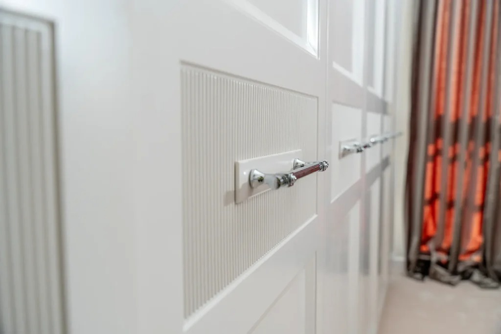 Biała szafa wnękowa z frezowanymi frontami i chromowanymi uchwytami, widok z boku, z zasłonami w tle. Detal wykończenia i elegancja w projekcie.