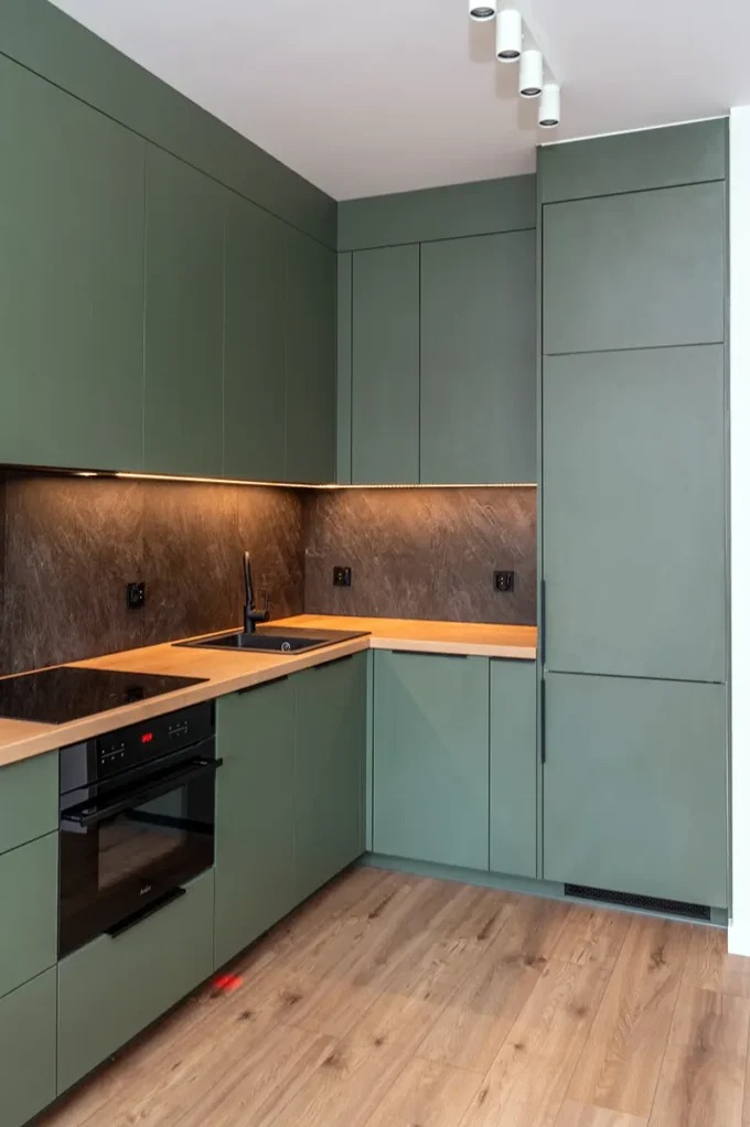 Kompaktowa kuchnia w odcieniach oliwki, z drewnianym blatem i czarnymi akcentami, oświetlona listwą LED. Nowoczesny minimalizm z widoczną płytą indukcyjną i piekarnikiem.