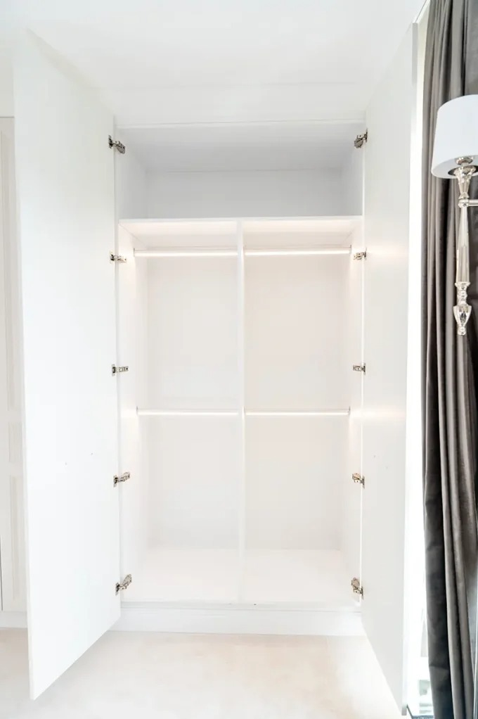 Otwarta, pusta, biała szafa wnękowa z półkami i drążkami, widoczne zawiasy. Obok szafy zasłona i lampa podłogowa w stylu glamour.