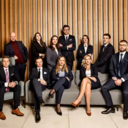 Zgrany zespół prawników z Krakowa w eleganckich strojach pozuje na tle drewnianej ściany, tworząc profesjonalny wizerunek kancelarii.