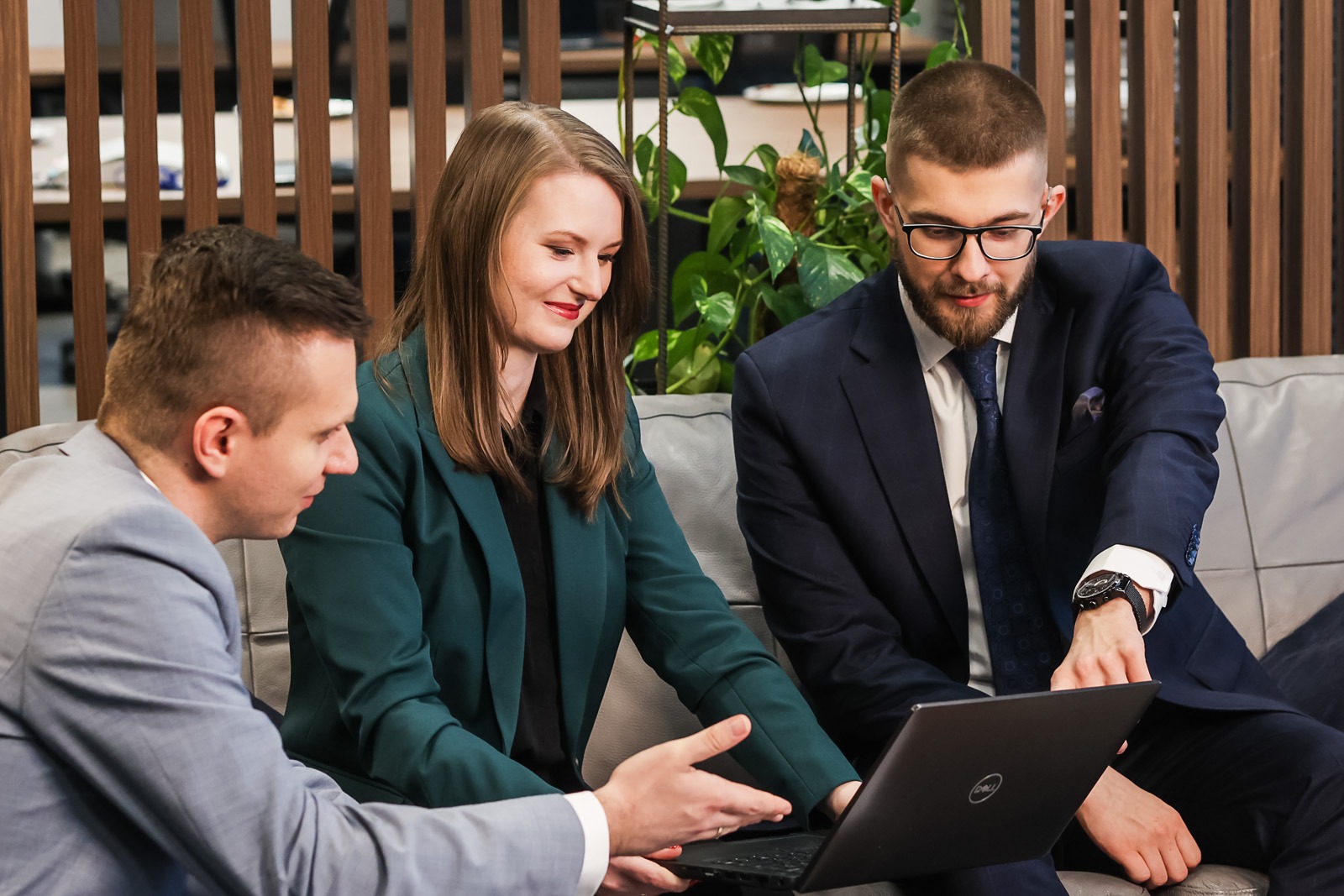 Trzech prawników w biurze analizuje dokumenty na laptopie. Kobieta w zielonej marynarce wskazuje na ekran, mężczyźni w garniturach obserwują. Neutralne tło, nowoczesny wystrój.
