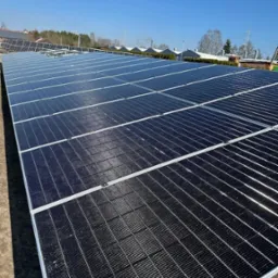 Rozległa instalacja fotowoltaiczna z widocznymi rzędami paneli, zamontowana na ziemi przy słonecznej pogodzie i błękitnym niebie. W tle budynki i drzewa.