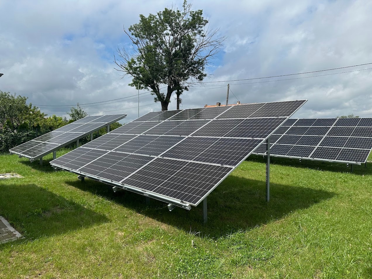 Instalacja paneli słonecznych na trawniku w słoneczny dzień. Widoczne panele, drzewo w tle i fragment budynku. Ujęcie z niskiej perspektywy.