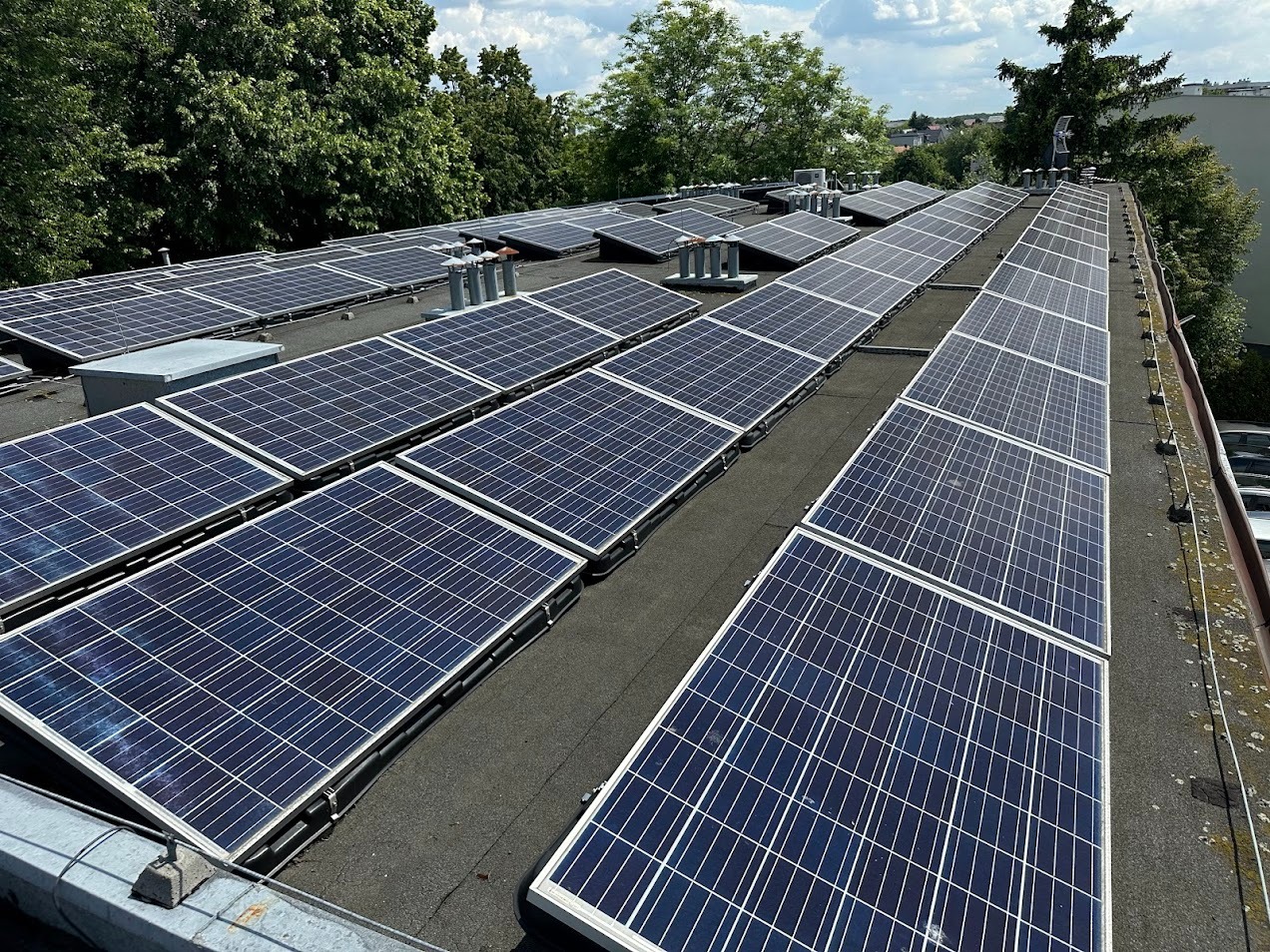 Rząd paneli słonecznych na płaskim dachu budynku, widok z góry, z kominami wentylacyjnymi i zielenią w tle. Układ paneli w rzędach, równolegle do krawędzi dachu.