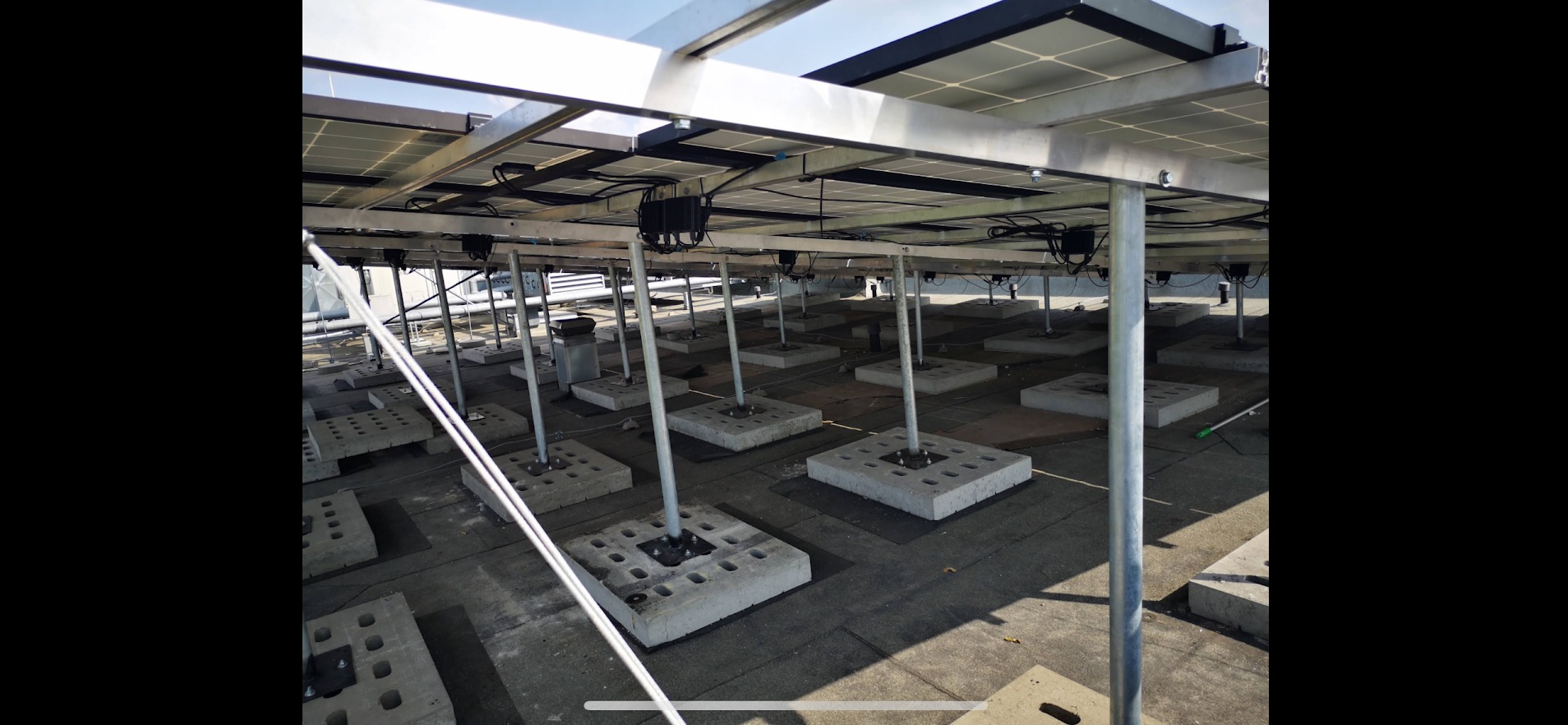 Montaż paneli słonecznych na płaskim dachu, widok od spodu. Stalowe podpory i betonowe podstawy. Widoczne kable i elementy konstrukcyjne.