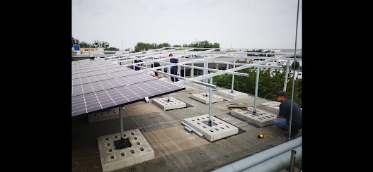 Montaż paneli na dachu budynku. Widoczne betonowe podstawy, stalowa konstrukcja wsporcza i częściowo ułożone panele. Prace instalacyjne w toku, dwóch monterów przy pracy.