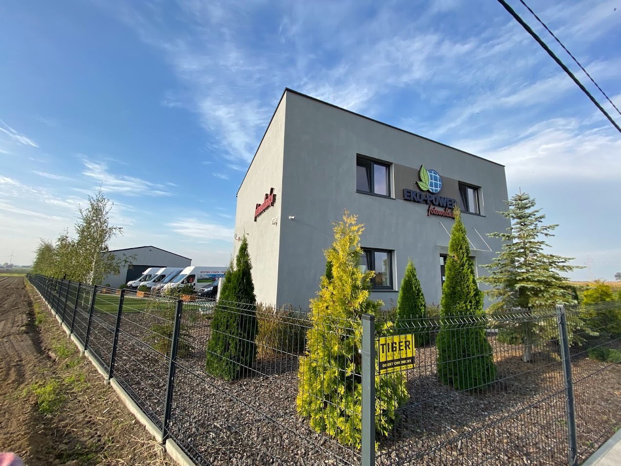 Nowoczesny budynek firmy Eko-Power Fotowoltaika z logo, otoczony zielenią i ogrodzeniem. Na parkingu zaparkowane białe busy. Słoneczny dzień.