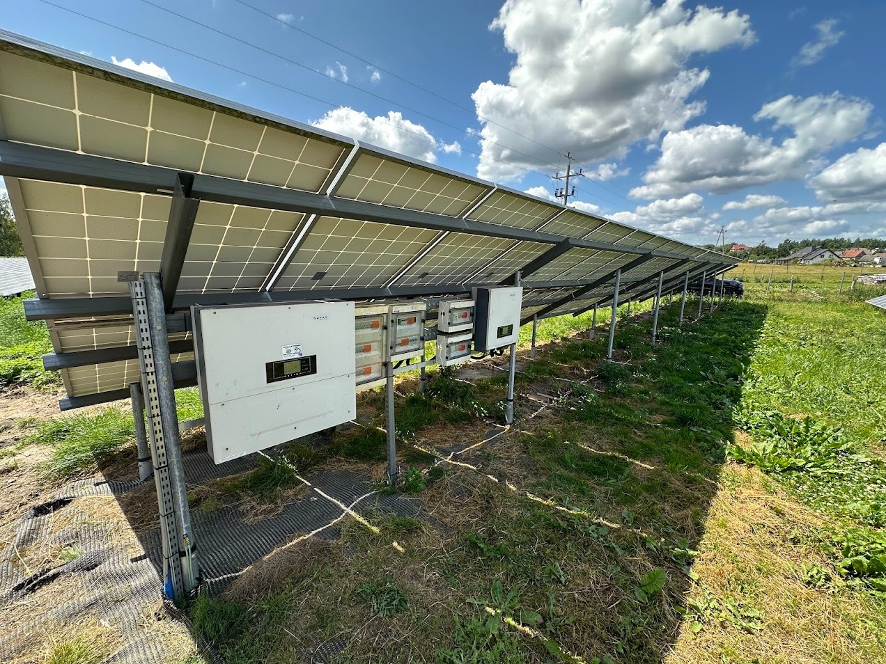 Rząd paneli słonecznych z widocznymi inwerterami na łące, w tle domy i błękitne niebo z chmurami. Widok z dołu, podkreślający perspektywę instalacji.