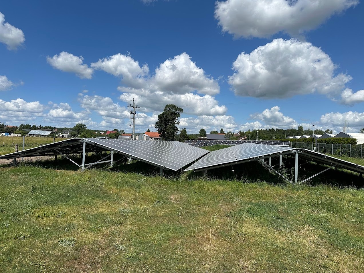 Rozległa instalacja paneli słonecznych na łące, z widokiem na wieś i błękitne niebo z chmurami. Panele ustawione w rzędach, wsparte na metalowych konstrukcjach.