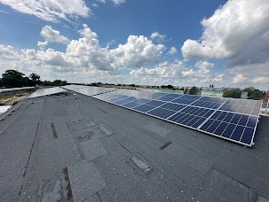 Panele słoneczne na płaskim dachu budynku w słoneczny dzień. Widoczne niebo z chmurami i fragment zabudowy miejskiej w tle. Perspektywa z góry.