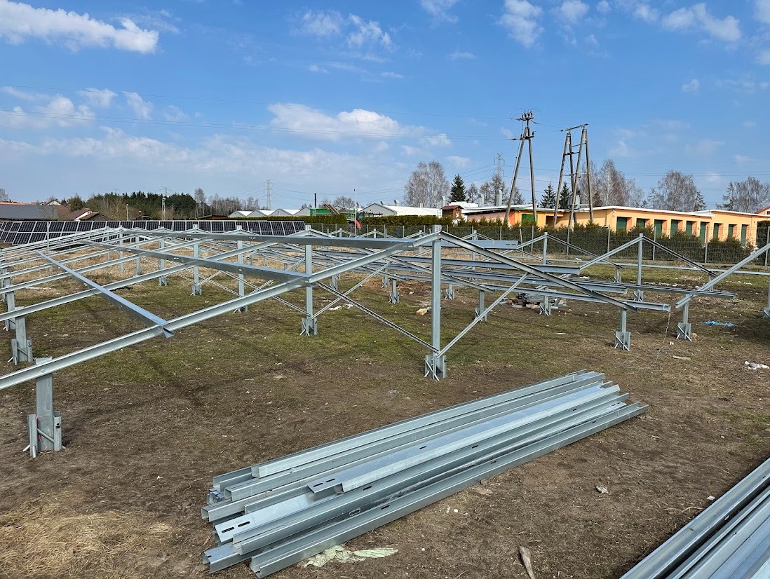 Montaż stalowej konstrukcji pod panele słoneczne na działce. W tle budynki i słupy energetyczne. Częściowo zamontowane panele widoczne z tyłu.