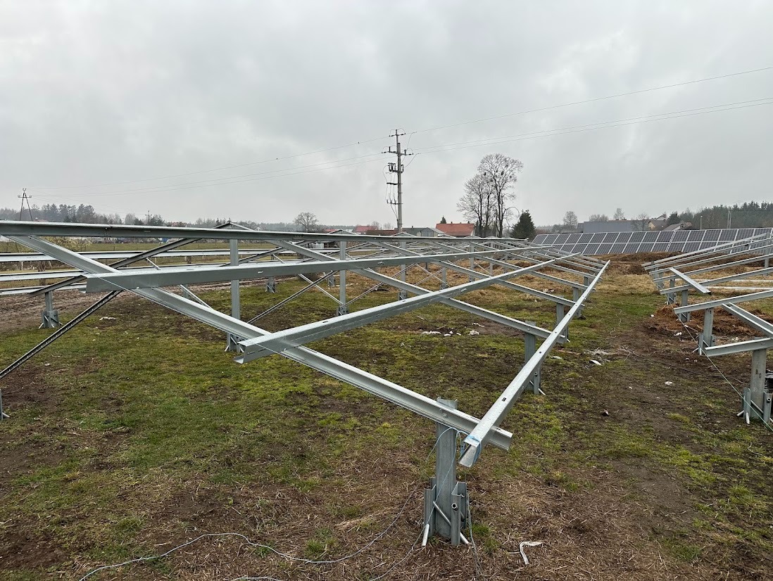 Montaż stelażu pod panele fotowoltaiczne na trawiastym terenie. Widoczne metalowe konstrukcje i częściowo zamontowane panele w tle. Pochmurne niebo.