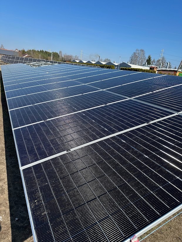 Rozległa instalacja fotowoltaiczna z widocznymi rzędami paneli, zamontowana na ziemi przy słonecznej pogodzie i błękitnym niebie. W tle budynki i drzewa.