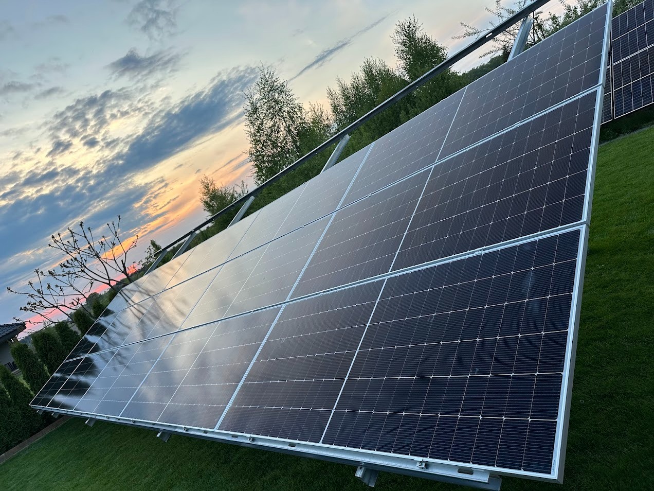 Panel słoneczny z widocznym odbiciem zachodzącego słońca na tle zielonego trawnika i drzew, ukazany pod kątem.