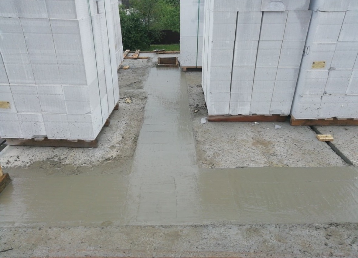 Wylany beton na fundamencie pod budowę. Widoczne białe bloczki betonowe na paletach. Świeży beton tworzy szarą, mokrą powierzchnię. Tło z zielenią.