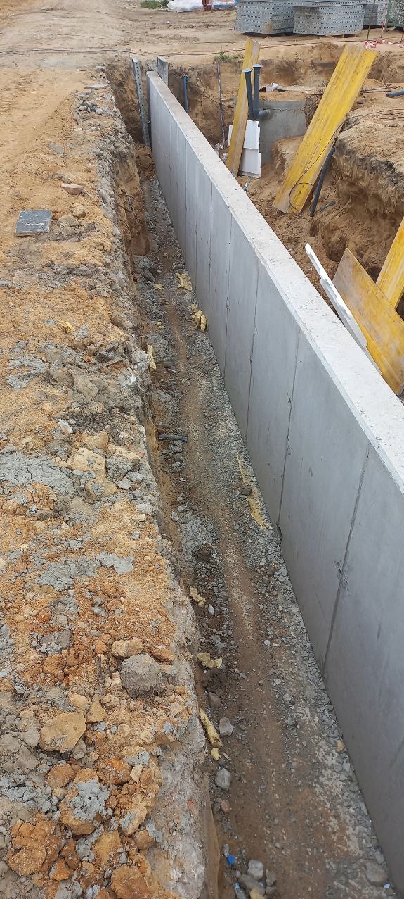 Betonowa ściana fundamentowa w wykopie. Widoczne zbrojenie i elementy konstrukcyjne. Żółte podpory i rury w tle. Grunt wokół wykopu.