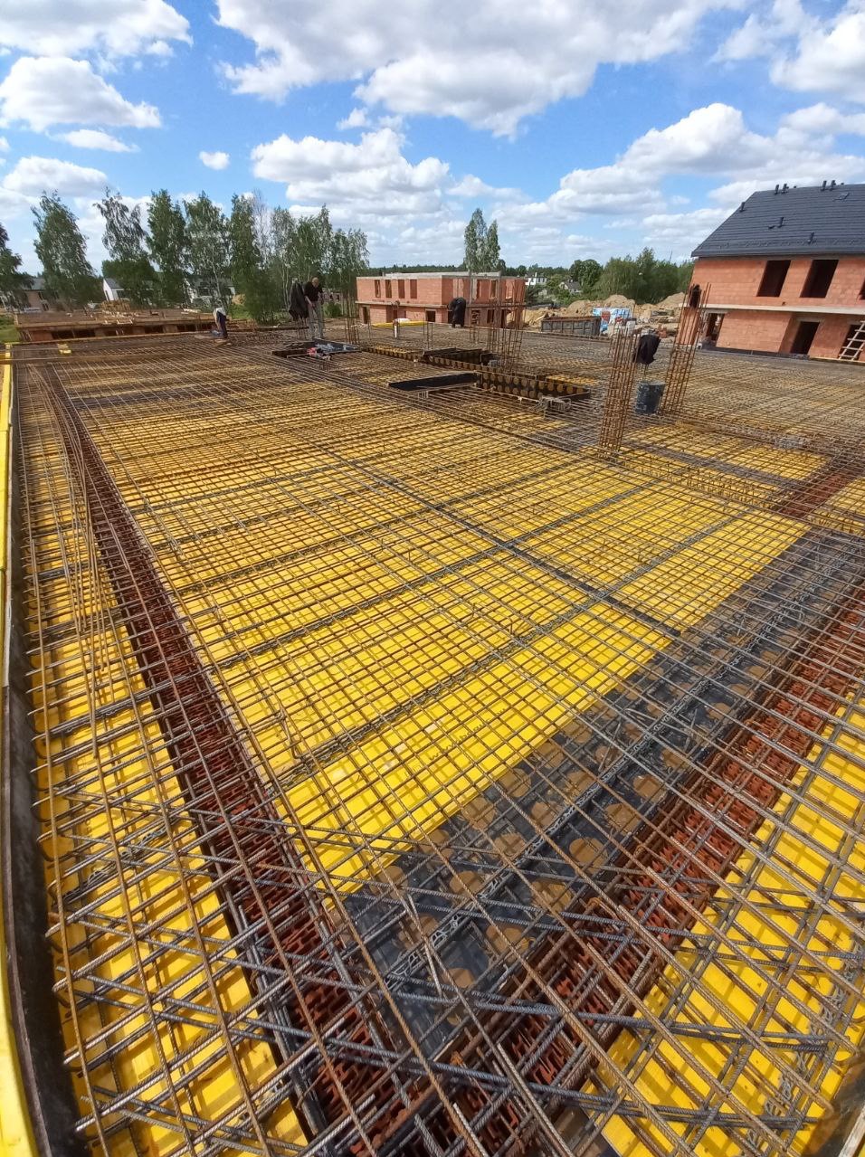 Zbrojenie stropu na żółtym szalunku, przygotowane do zalania betonem. W tle budynek w stanie surowym otwartym i pracujący robotnicy.