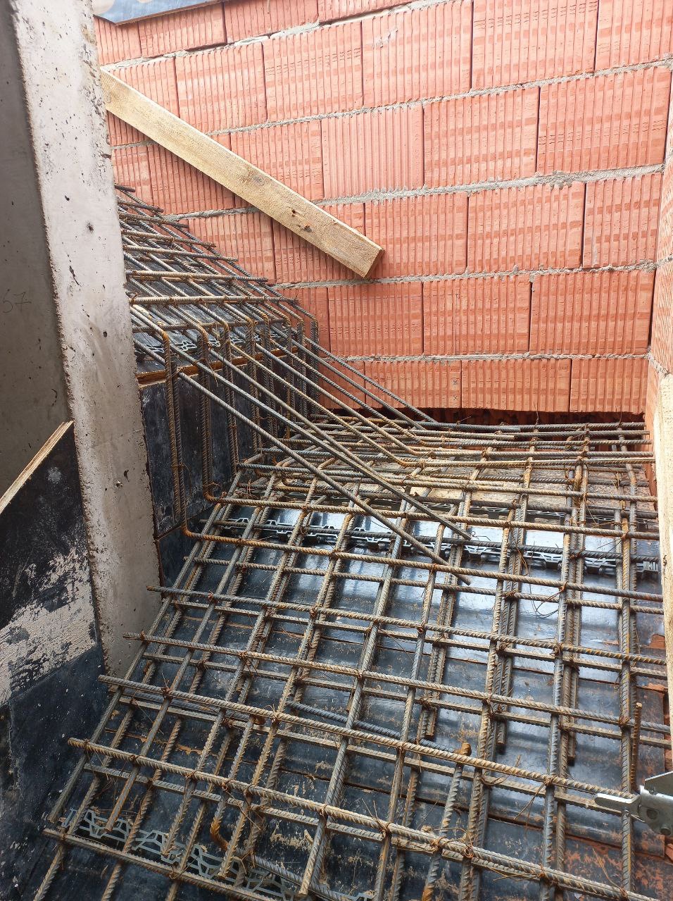 Zbrojenie schodów z ceglanymi ścianami w tle. Widoczna drewniana listwa. Detal budowy domu z żelbetu, z bliska. Metalowe pręty tworzą siatkę.