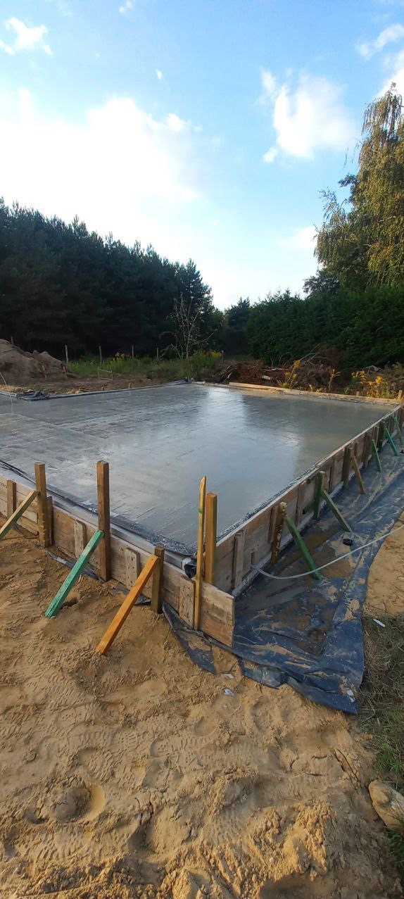 Wylana, gładka płyta fundamentowa otoczona drewnianym szalunkiem na piaszczystym terenie budowy, z widoczną folią hydroizolacyjną i zielenią w tle.