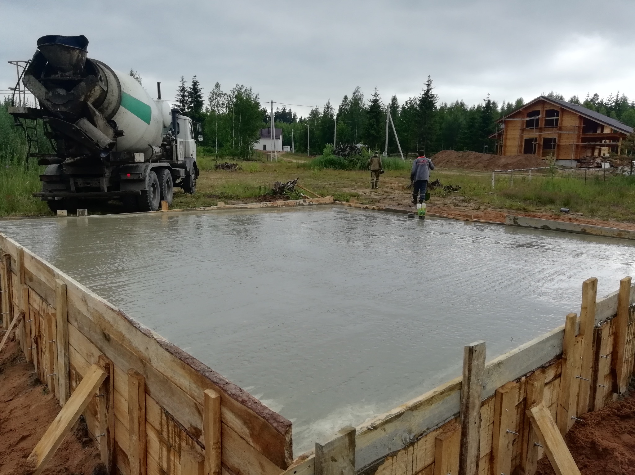 Zalany fundament pod budowę domu jednorodzinnego z drewnianą konstrukcją szalunkową. W tle betoniarka i pracownicy na tle lasu.