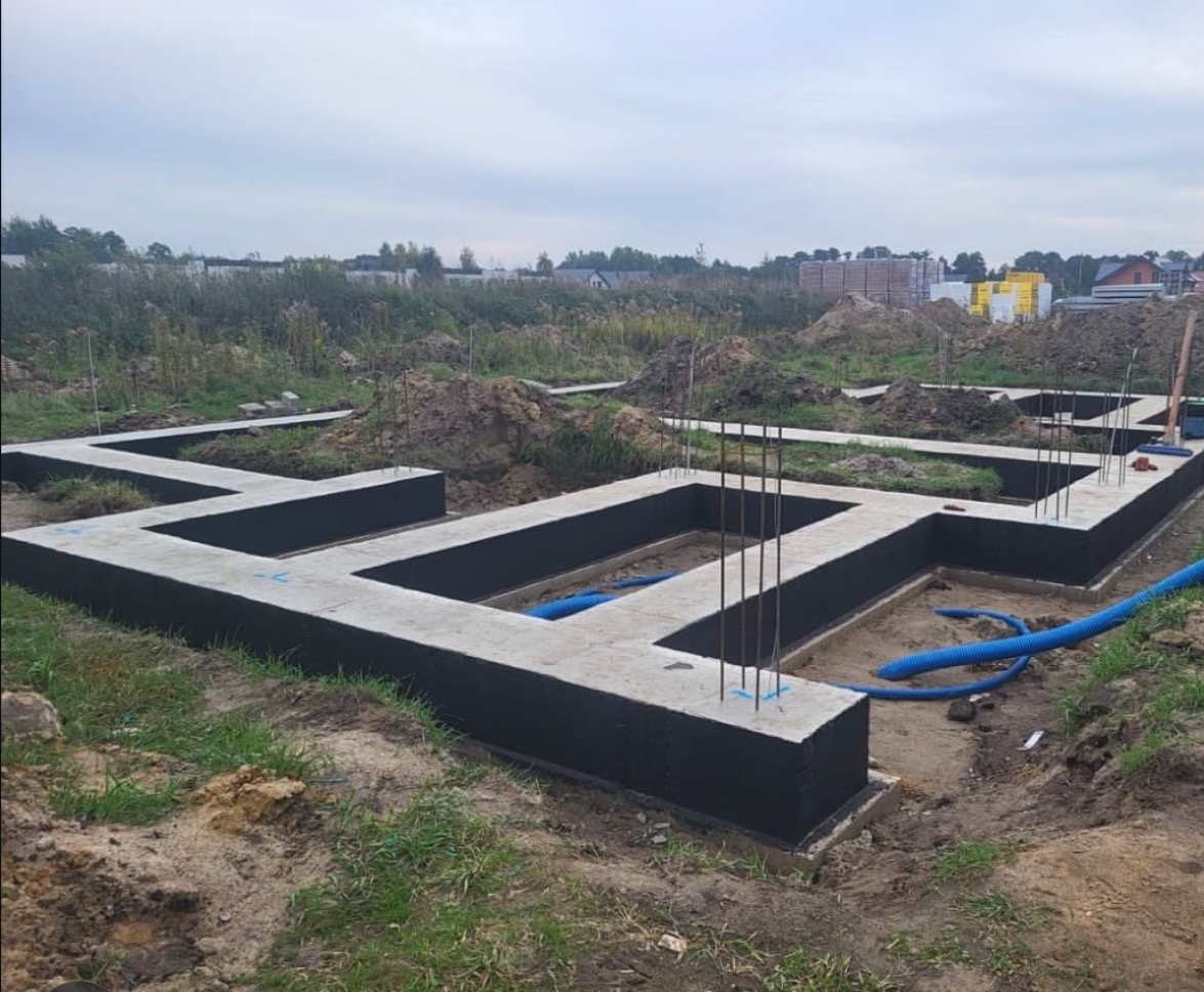 Zalane fundamenty domu z hydroizolacją, wystające pręty zbrojeniowe, niebieskie rury wewnątrz, ziemia i trawa w tle. Widok z poziomu gruntu.