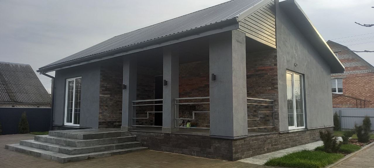 Parterowy dom z szarą elewacją, ceglanym wykończeniem i metalowym dachem, widok z zewnątrz w pochmurny dzień. Widoczne schody wejściowe i fragment ogrodu.