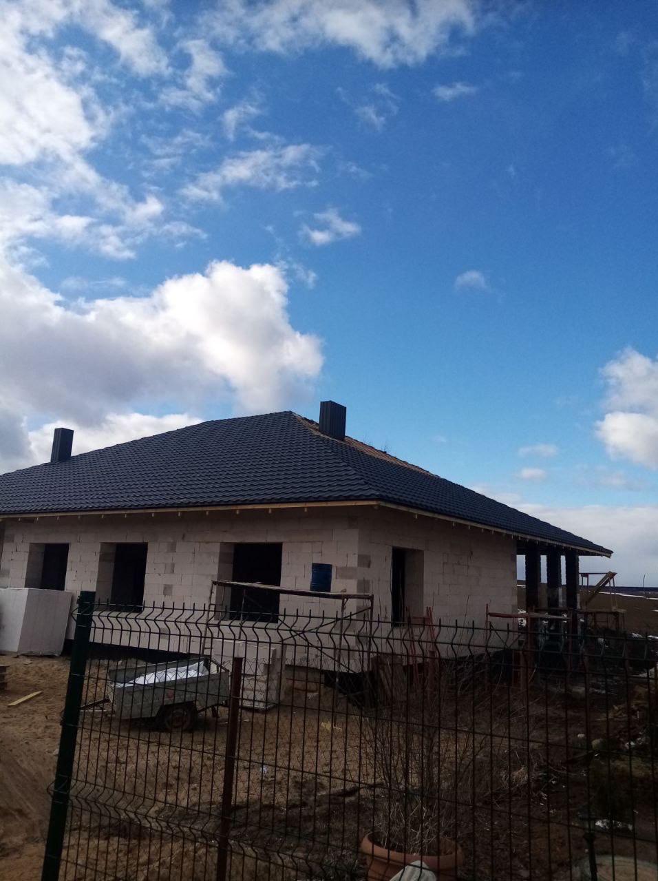 Surowy dom z czarnym dachem z dachówki, widoczny przez ogrodzenie. Ściany z białych bloczków, otwory okienne i drzwiowe. Błękitne niebo z chmurami w tle.