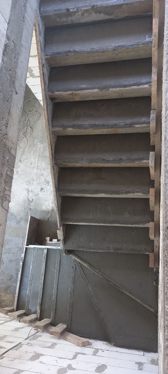 Betonowe schody w trakcie budowy, widok z dołu, szalunek drewniany częściowo widoczny, surowy stan, białe kable zwisają ze ściany.