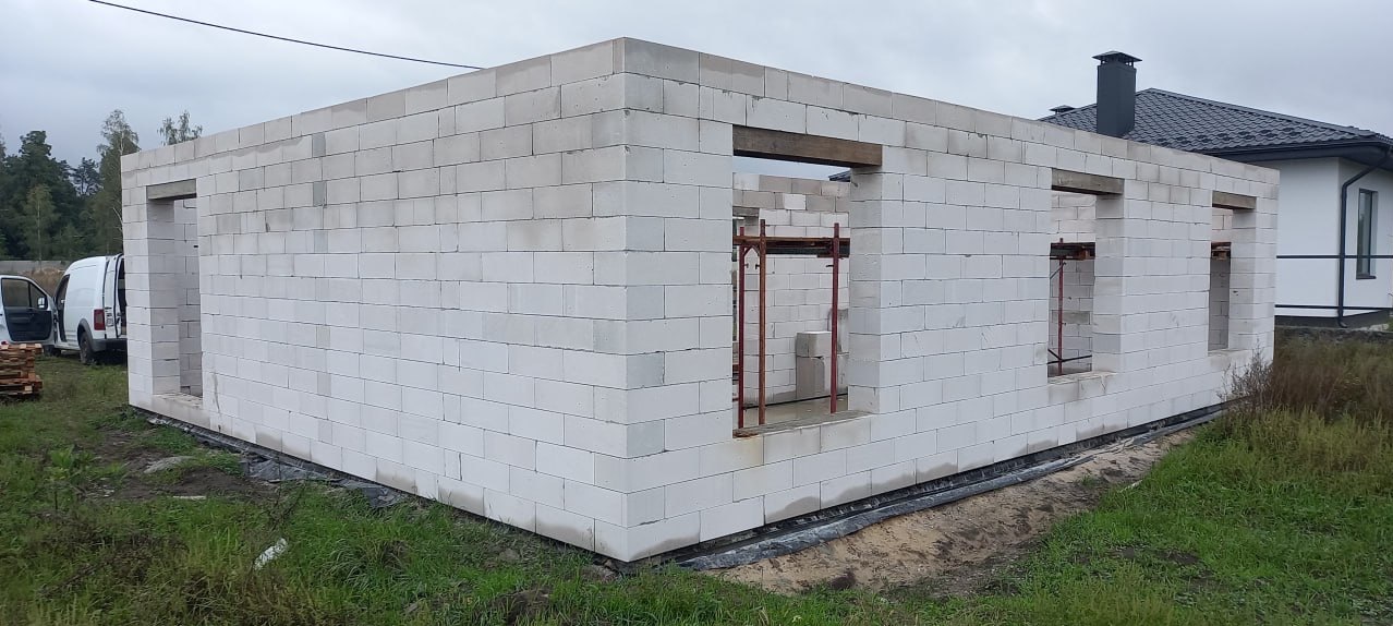 Surowy stan domu z białych bloczków, widoczne otwory okienne i drzwiowe z drewnianymi belkami. Na zewnątrz trawa i częściowo widoczny budynek z czarnym dachem.