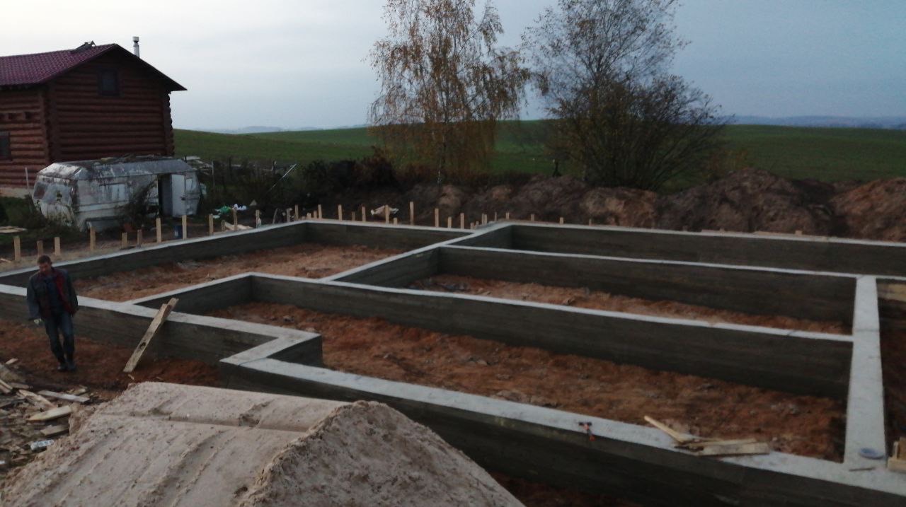 Zalane fundamenty pod budynek na działce z przyczepą kempingową i drewnianym domkiem w tle, widok z góry, pochmurny dzień.