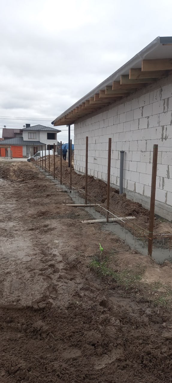 Budowa domu: fundamenty zbrojone stalą, ściany z białego betonu komórkowego. Widoczne drewniane elementy konstrukcji dachu i osoba w tle z parasolem.