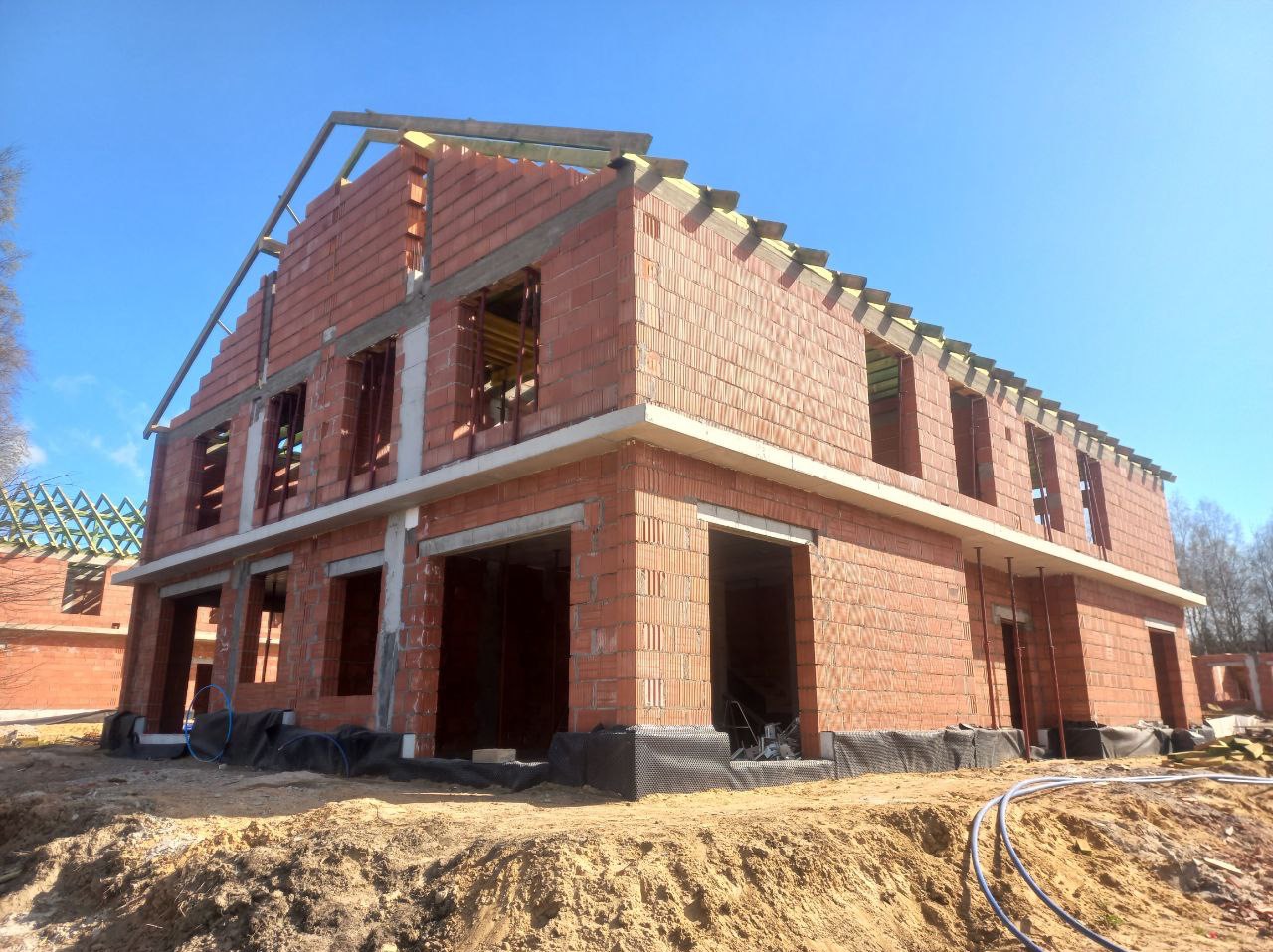 Dwukondygnacyjny dom w trakcie budowy z czerwonej cegły, widoczne otwory okienne i zarys dachu. Grunt piaszczysty, niebieskie niebo w tle.