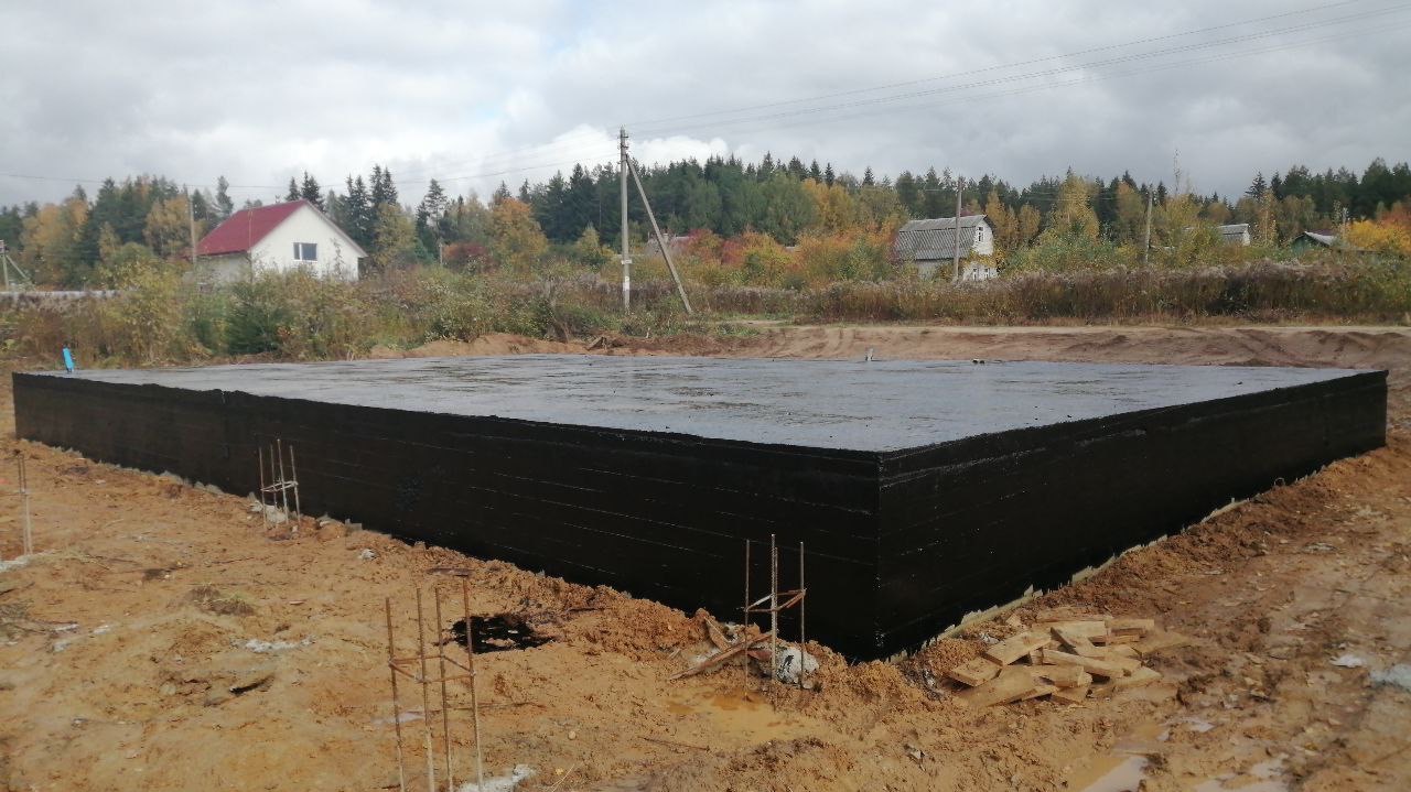 Zabezpieczony, czarny fundament płyty budynku na gruncie z wystającymi prętami zbrojeniowymi, w tle zabudowa jednorodzinna i jesienne drzewa.