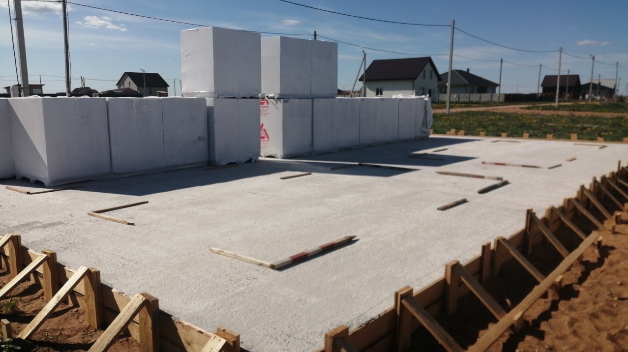 Zalany fundament z drewnianym szalunkiem i bloczkami Ytong w tle. Widoczne domy w oddali. Słoneczny dzień.