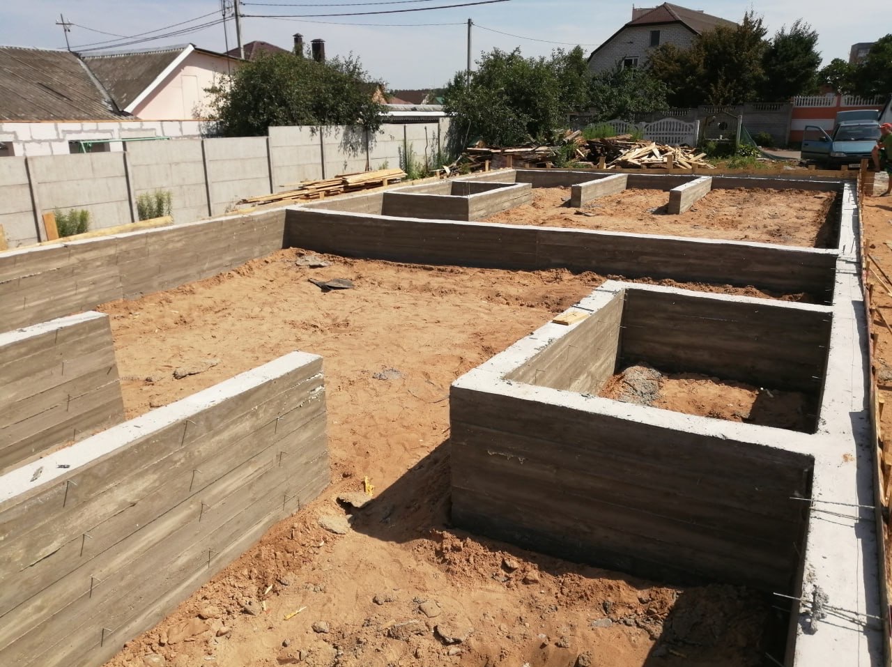 Zalane betonem fundamenty domu jednorodzinnego na piaszczystej działce, widoczne zbrojenia i deski szalunkowe, w tle ogrodzenie z prefabrykatów betonowych.