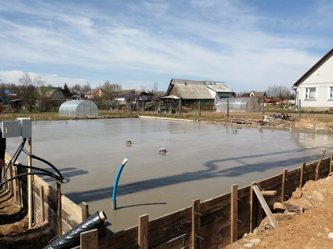 Zalany fundament pod budowę domu jednorodzinnego. Widoczne szalunki z desek, rury instalacyjne wystające z betonu. W tle zabudowa wiejska i szklarnie.
