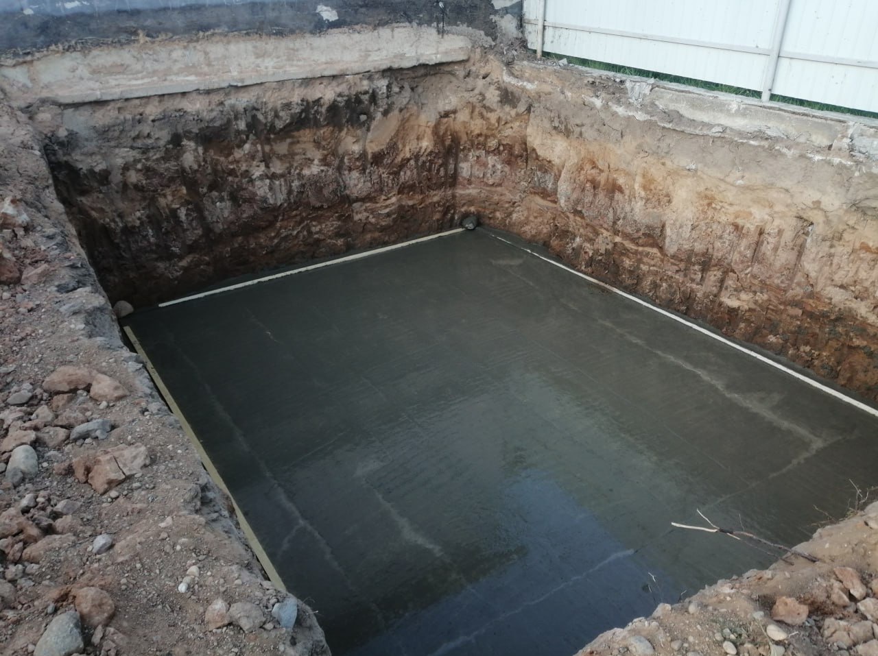 Wykop pod fundament z wylaną, świeżą betonową płytą. Widoczne ściany wykopu z warstwami ziemi i biała listwa wzdłuż krawędzi płyty. Górny kadr ukazuje fragment ogrodzenia.