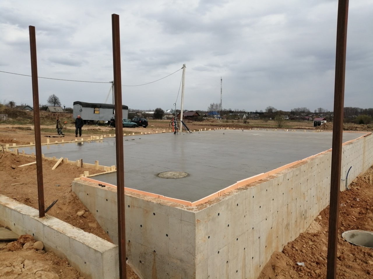 Wylany fundament pod budynek z widoczną izolacją termiczną i zbrojeniami, na tle pochmurnego nieba i zabudowań wiejskich. Prace betoniarskie w toku.