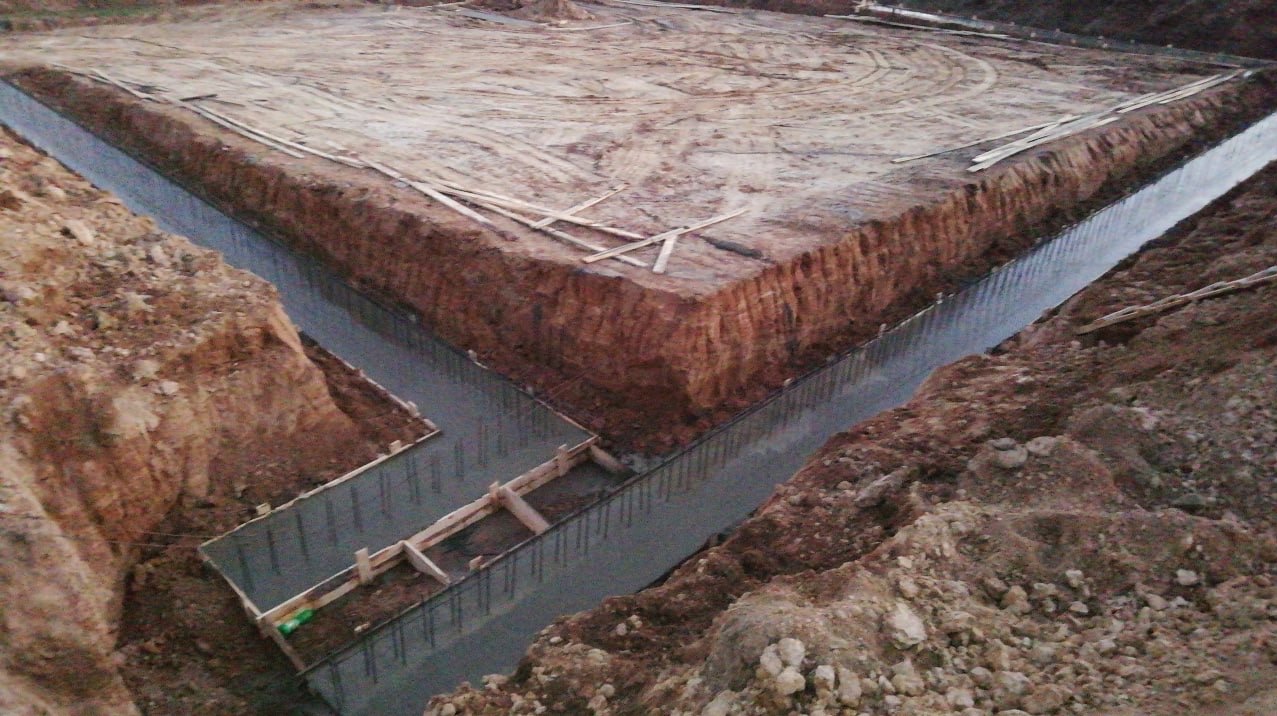 Zalane betonem fundamenty pod budowę domu, widoczne szalunki i wystające pręty zbrojeniowe. Wykopane rowy fundamentowe z ziemią wokół.