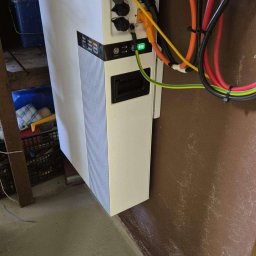 GROMAR-AUTO Mariusz Gromowski - Biały magazyn energii z podłączonymi kablami, widoczne diody LED i złącza. Instalacja w pomieszczeniu o brązowych ścianach i szarej podłodze.