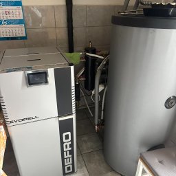 GROMAR-AUTO Mariusz Gromowski - Kompaktowy piec na pellet marki Defro Evopell obok szarego zasobnika ciepłej wody w kotłowni z kalendarzem na ścianie. Widoczne rury i instalacja.