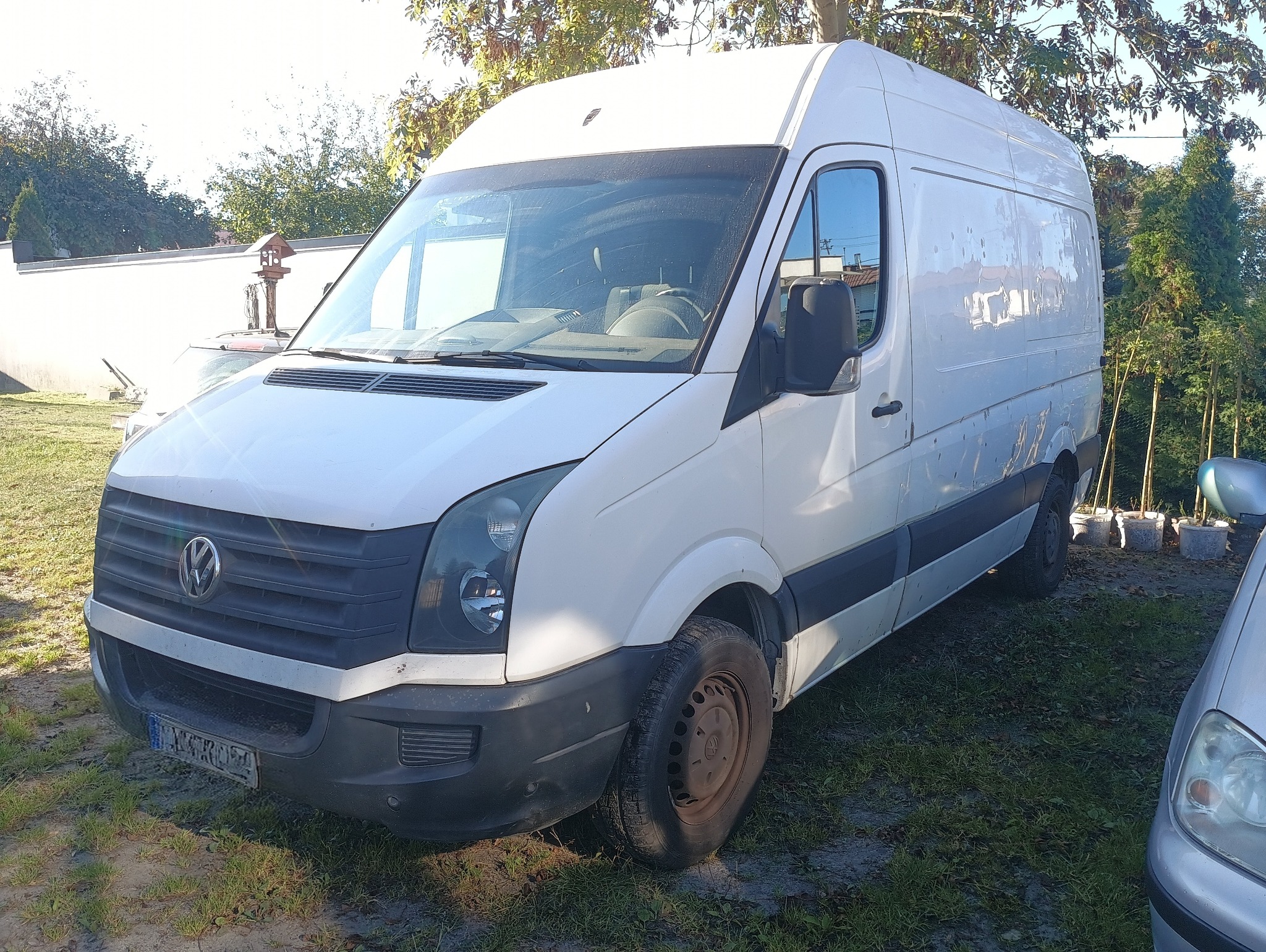 Biały bus Volkswagen Crafter na trawiastym podłożu, widok z przodu i boku, zarys drzew w tle. Pojazd gotowy do transportu lub przeprowadzki.