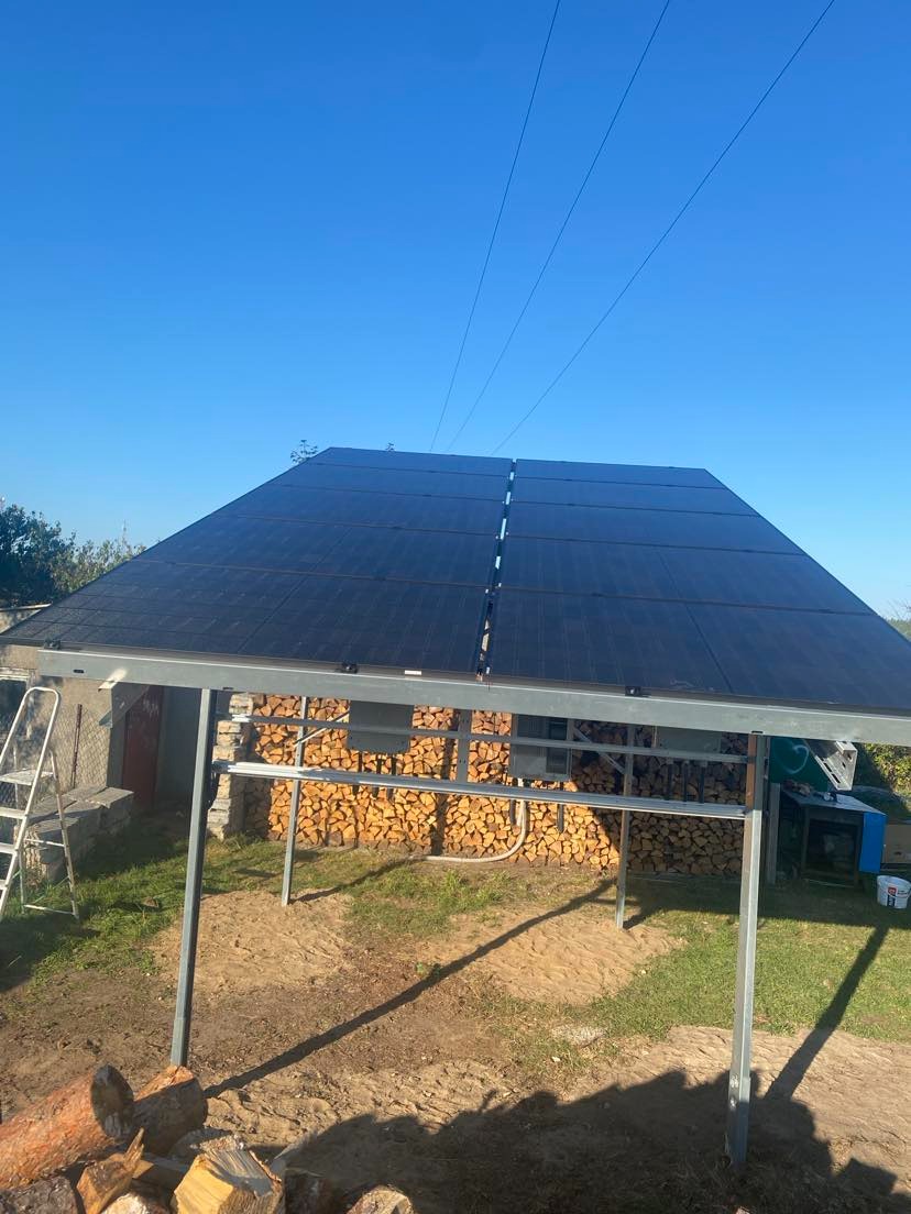 Zadaszona instalacja fotowoltaiczna z metalową konstrukcją, na tle ściany z ułożonym drewnem opałowym, w słoneczny dzień. Widoczne przewody elektryczne.