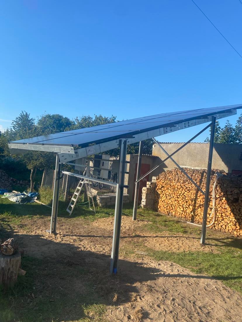 Montaż paneli fotowoltaicznych na metalowej konstrukcji w ogrodzie, widok na panele pod błękitnym niebem, w tle stos drewna.